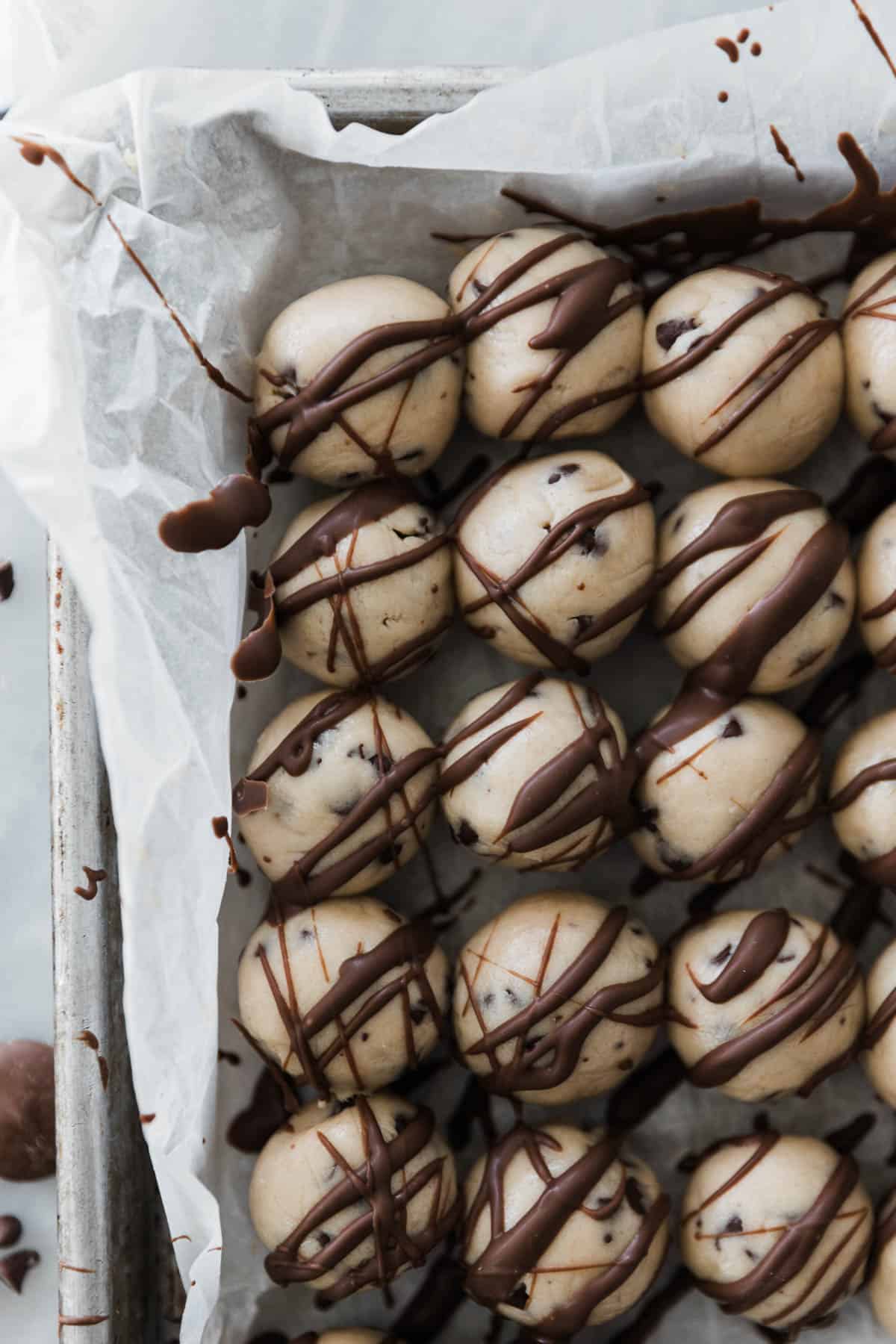 Several small cookie dough balls with chocolate coating drizzled on a baking sheet. 