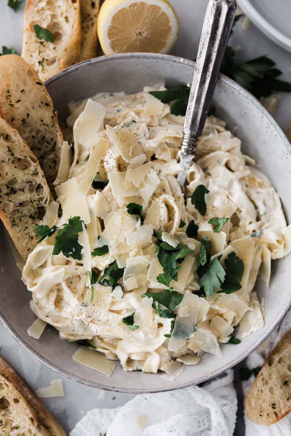 Homemade Alfredo sauce with fettuccine noodles topped with parsley in a bowl with breadsticks. 