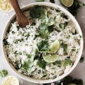 A big bowl of cilantro lime rice topped with limes and a wooden spoon.