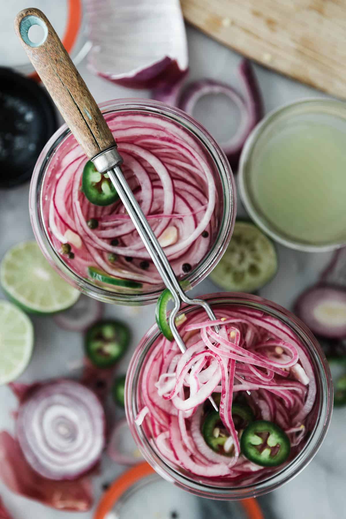 Two jars filled with sliced red onions vinegar, spices and sliced jalapeños with a large prong on top.