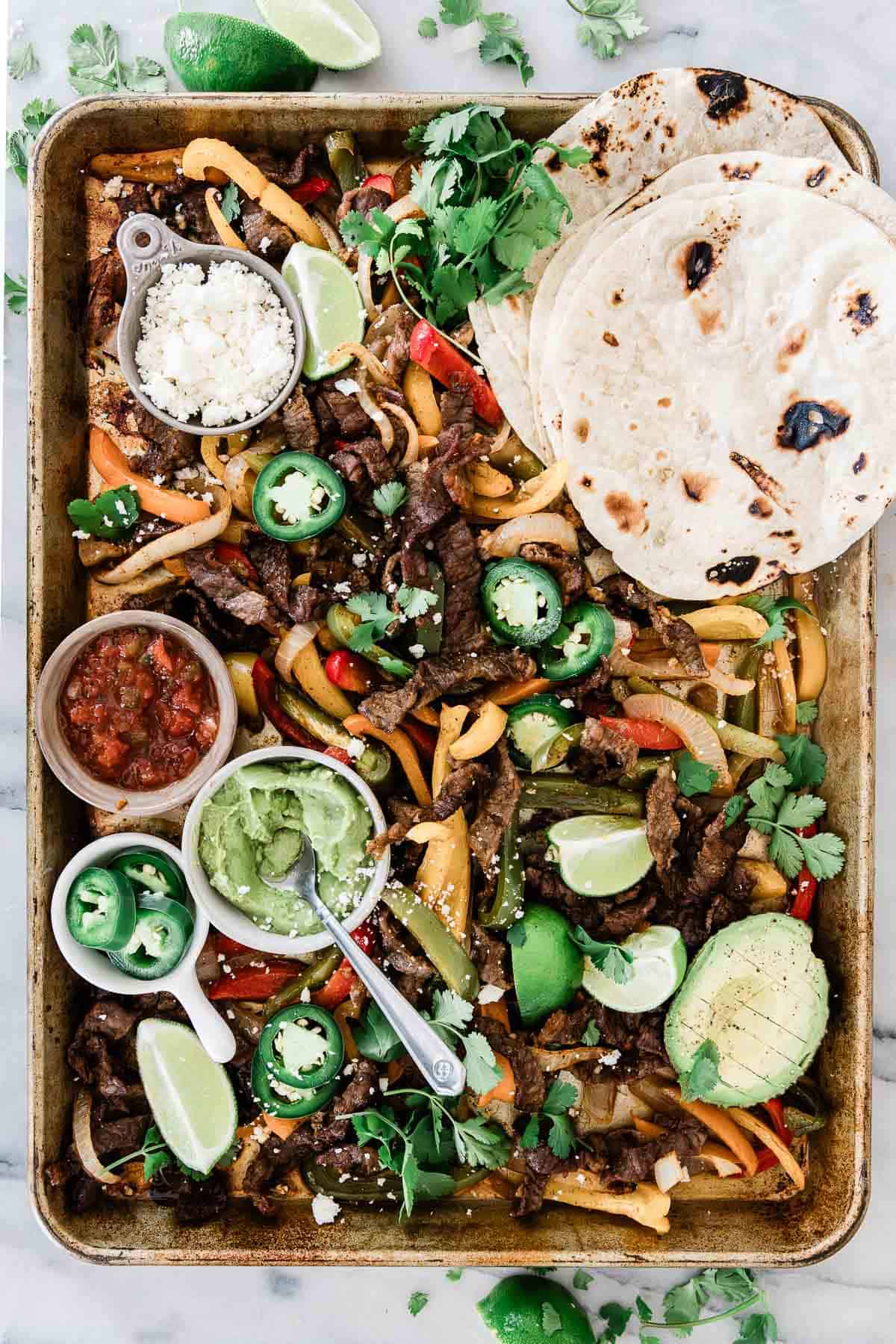 Sheet pan steak fajitas on a large pan. There are tortillas in one corner and condiments in the opposite corner.