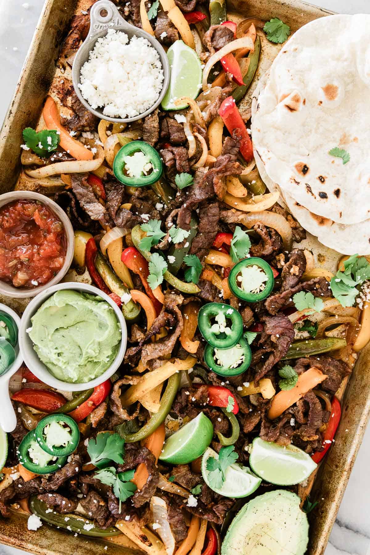 Sheet pan steak fajitas on a sheet tray. There are tortillas, condiments, limes, and jalapeños also on the tray.