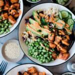 Salmon salad bowl in white bowls with black rims. There are small bowls of cucumber and edamame to the side,