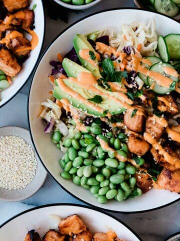 Salmon salad bowl in white bowls with black rims. There are small bowls of cucumber and edamame to the side,
