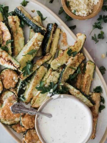 A platter of air fryer zucchini with a bowl of parmesan cheese on the side and a creamy white sauce.