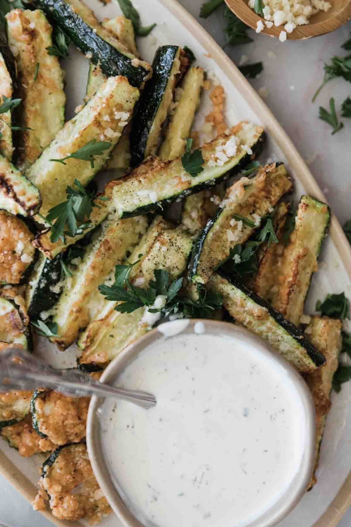Air fried zucchini on a platter with a bowl of creamy dip.