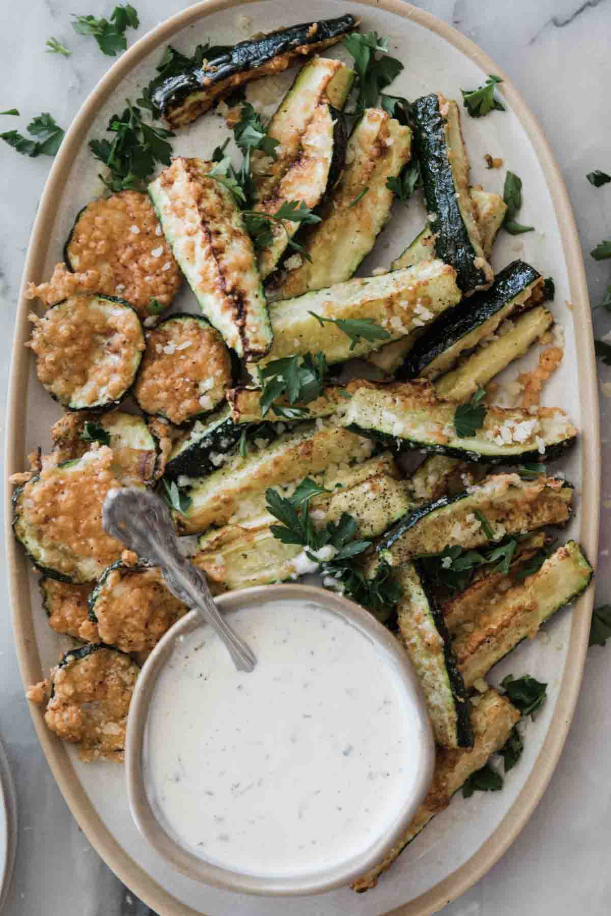 A platter of air fryer zucchini with a bowl of white sauce with a spoon in it.