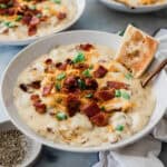 A bowl of loaded baked potato soup on the table topped with bacon, cheese, and green onions.