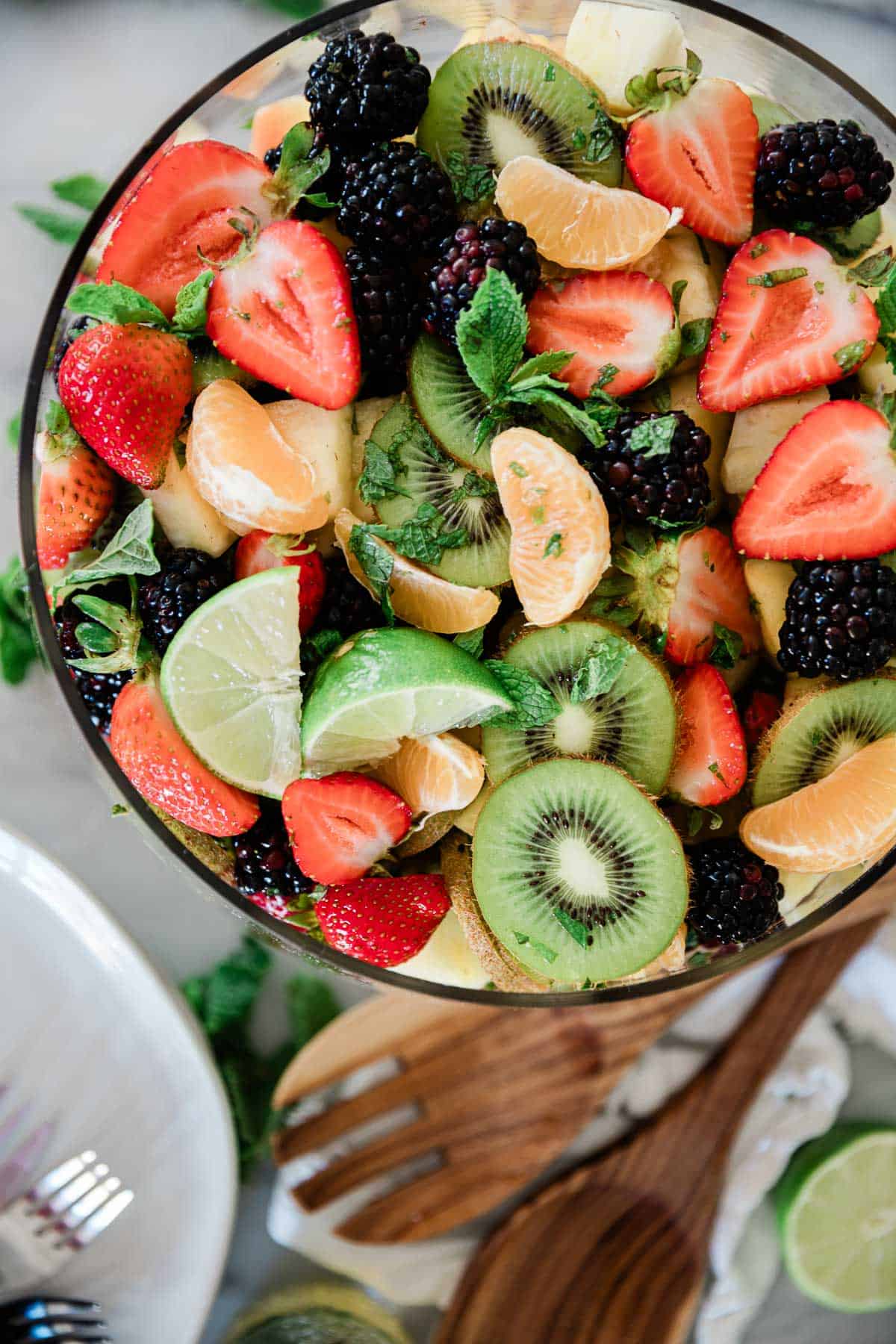 A bowl of fresh fruit salad on a table with a spoon and plates to the side.