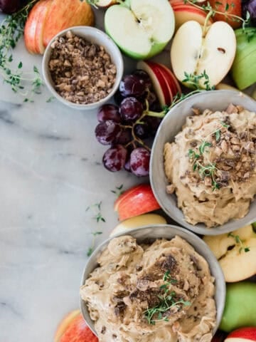 Bowls of cream cheese apple dip on a marble block with fruit and nuts.