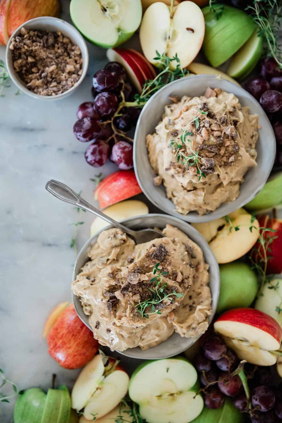 Two bowls of creamy apple dip on a table with grapes and apples around them.