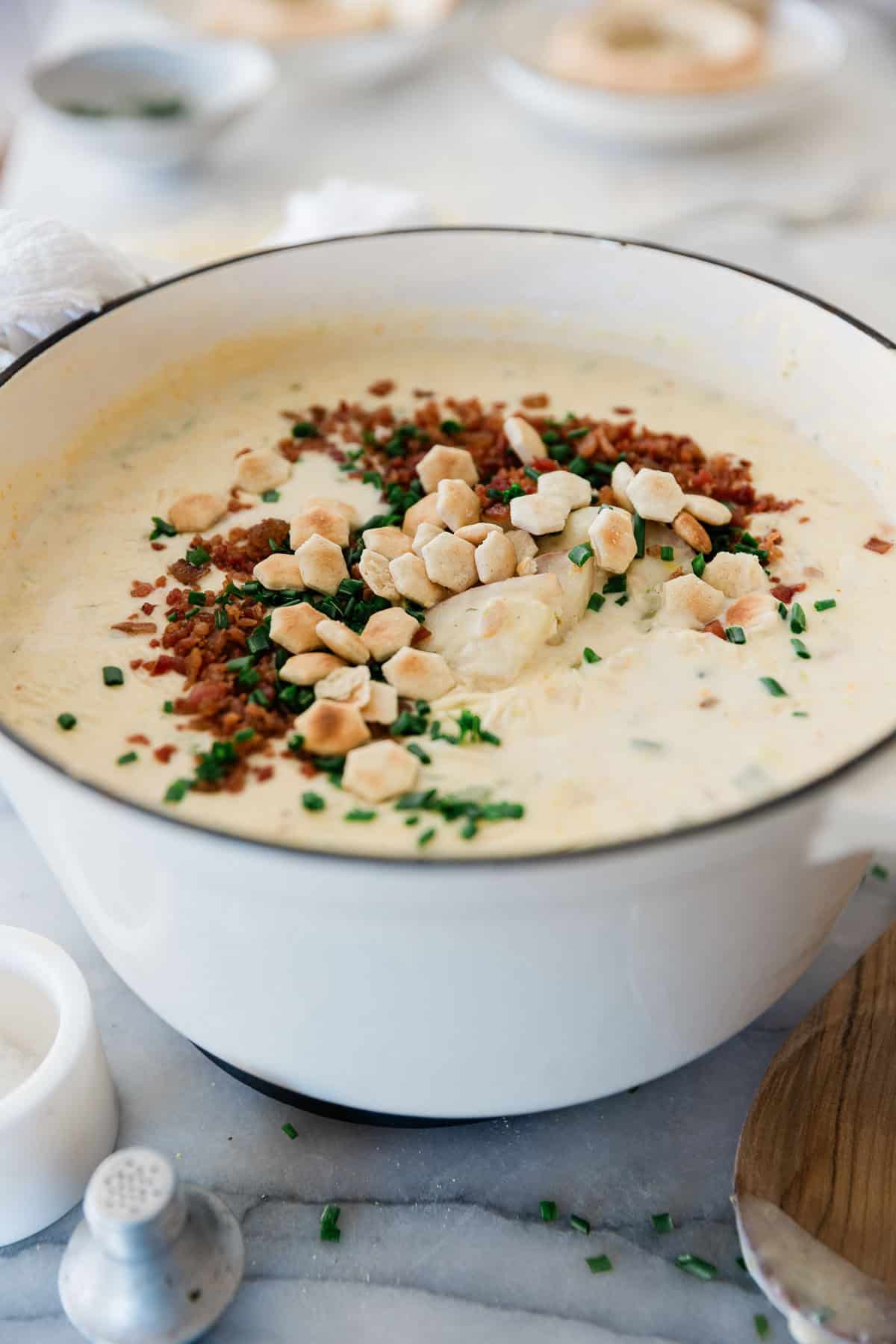 Boston clam chowder in a pot garnished with oyster crackers, bacon bits, and chopped parsley.