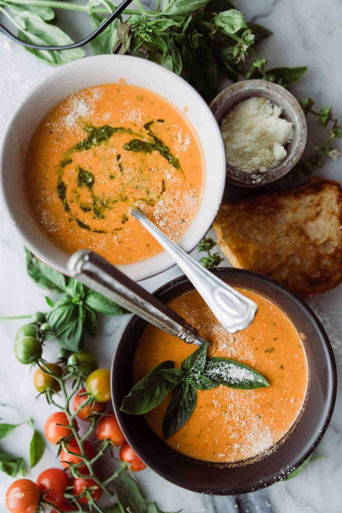Creamy tomato soup in white bowls that are black and white. They are garnished with basil and pesto.