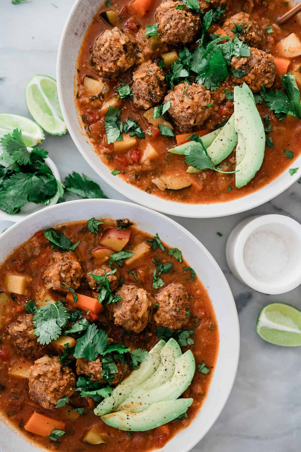 Two bowls of Mexican meatball soup on a table garnished with avocado and cilantro.