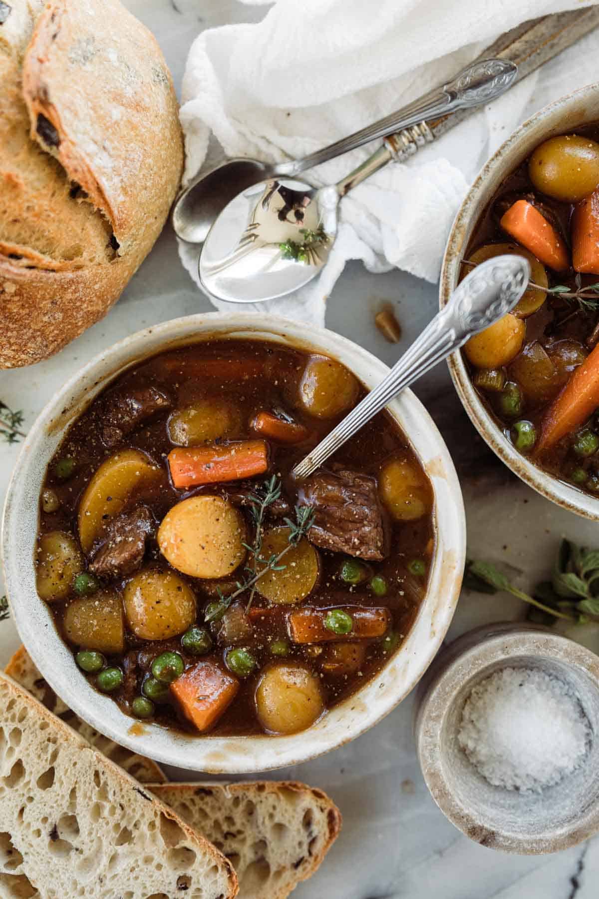 A bowl of homemade beef stew with bread on the table to the side. 