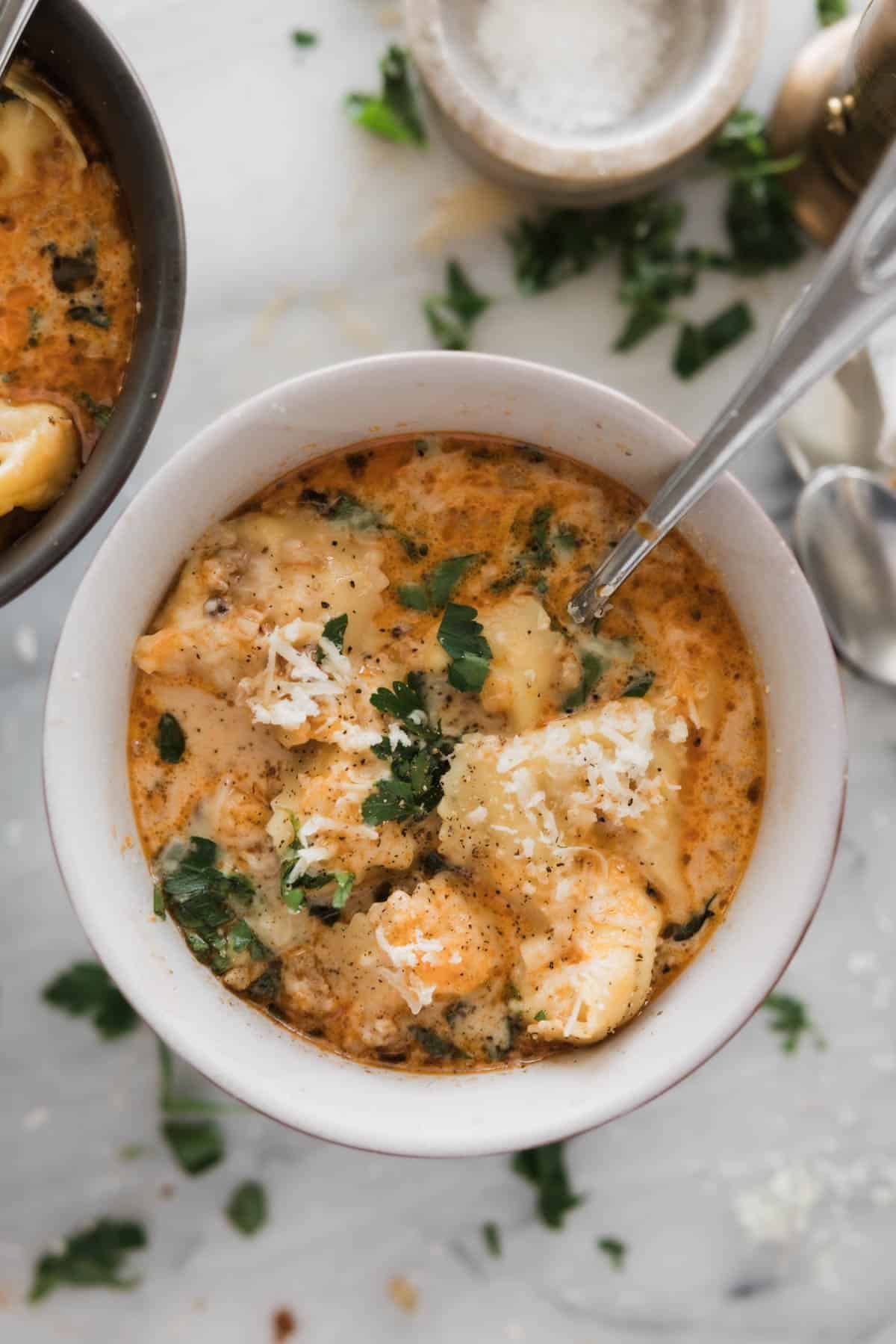A bowl of sausage tortellini soup on a table with herbs around it on the table. 