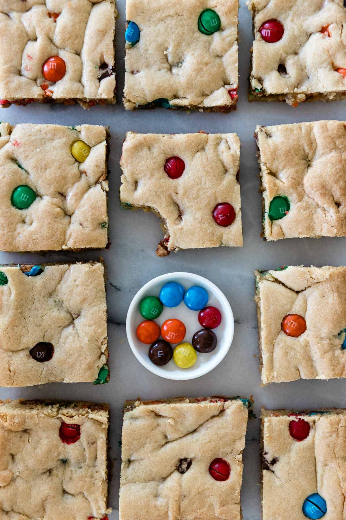 Homemade blondies cut into squares on a baking tray with a dish of M&M's on the  tray. 