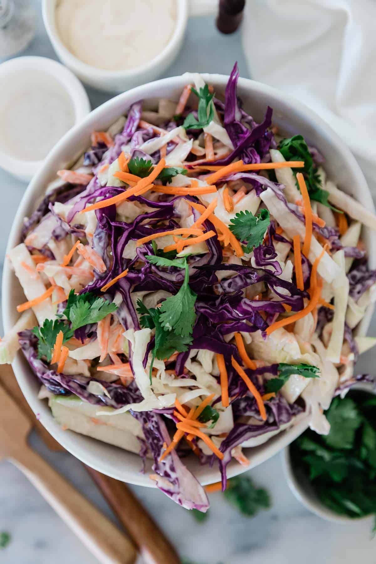 Coleslaw with apples in a bowl with a dish of more dressing and parsley on the side. 