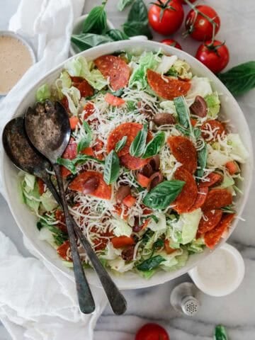 A bowl filled with salad with pepperoni and two spoons on the side.