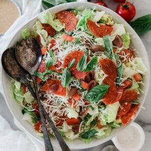 A bowl filled with salad with pepperoni and two spoons on the side.