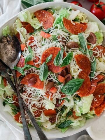A bowl filled with salad with pepperoni and two spoons on the side.